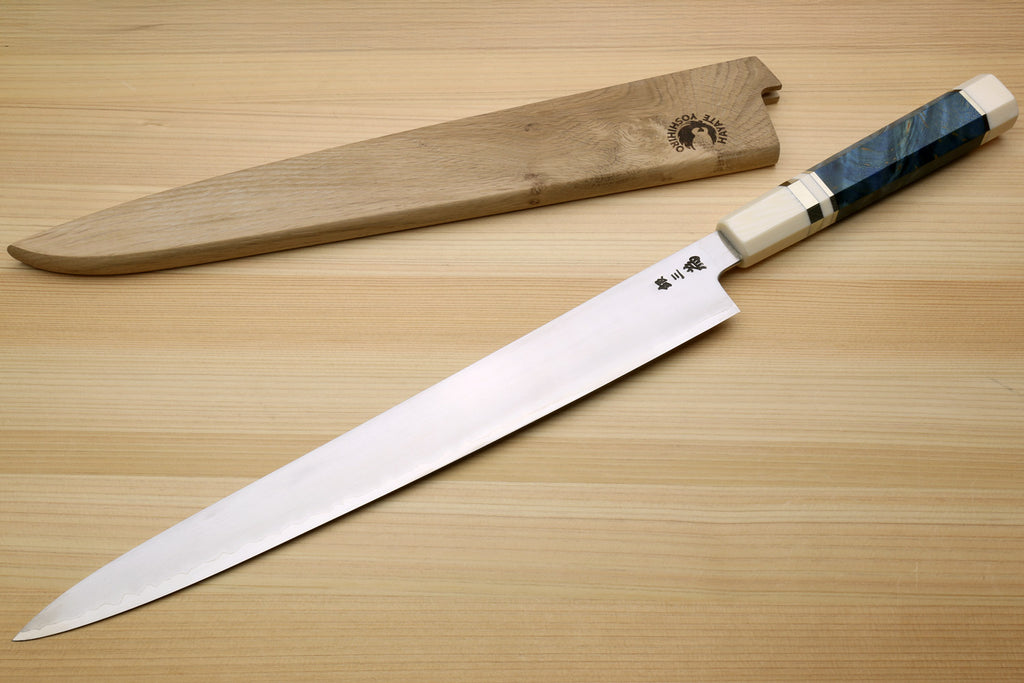 A Japanese-style kitchen knife with a long silver blade and a dark blue marbled handle, displayed diagonally next to its wooden sheath on a light wood grain surface.