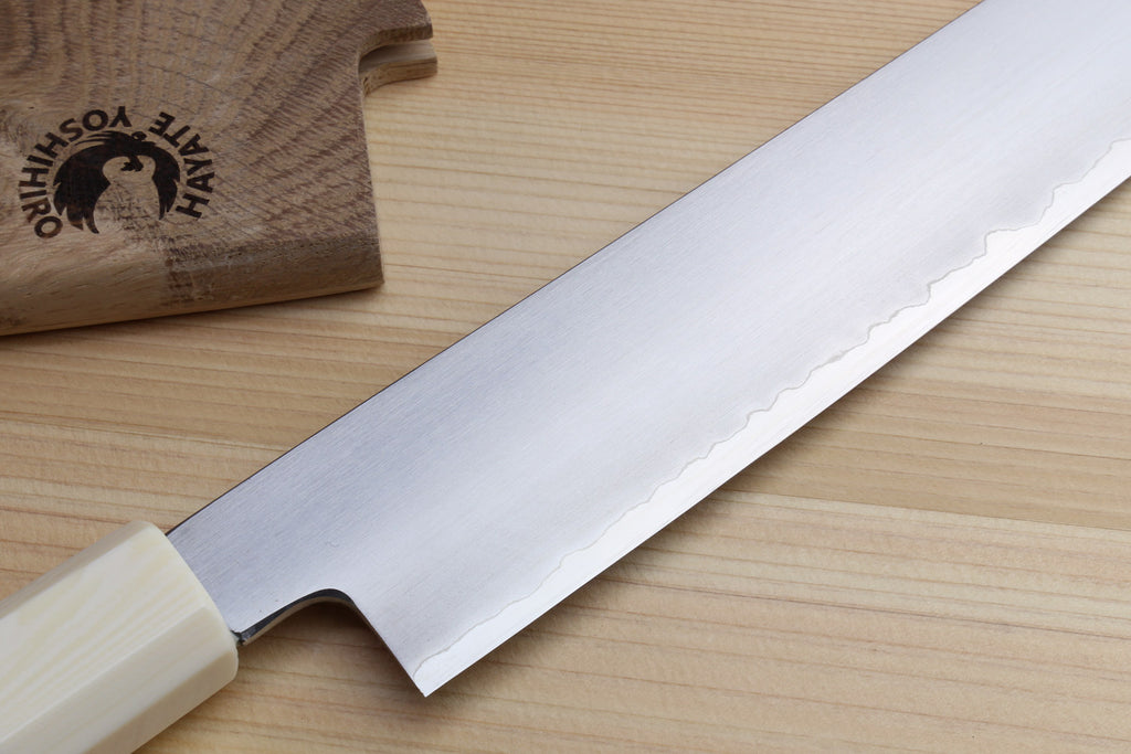 A Japanese-style kitchen knife with a satin-finished silver blade and a cream-colored handle, resting diagonally on a light wood surface next to a branded wooden block.