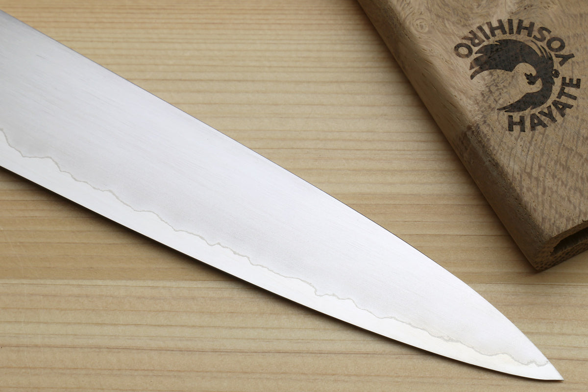 A close-up of a Japanese-style kitchen knife blade resting diagonally on a light wood surface, with part of a wooden box engraved with the Onihishima Hayate logo visible in the upper right.