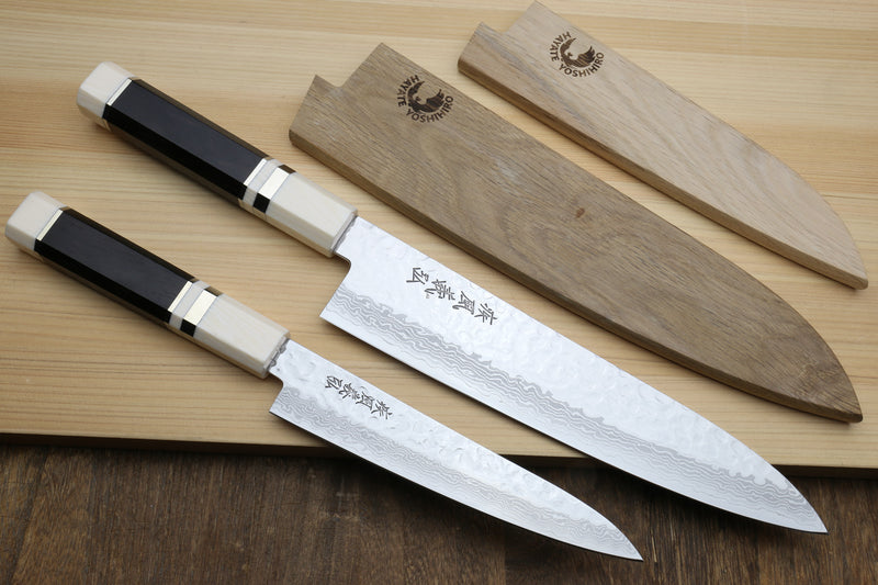Two Japanese kitchen knives with black and cream handles are displayed diagonally on a light wood cutting board next to their matching wooden sheaths. The blades feature a hammered finish and are engraved with Japanese characters.