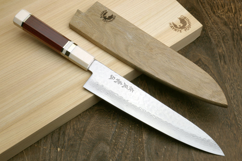 A Japanese-style kitchen knife with a patterned steel blade and a reddish-brown wooden handle, resting diagonally on a light wood cutting board next to its matching wooden sheath.