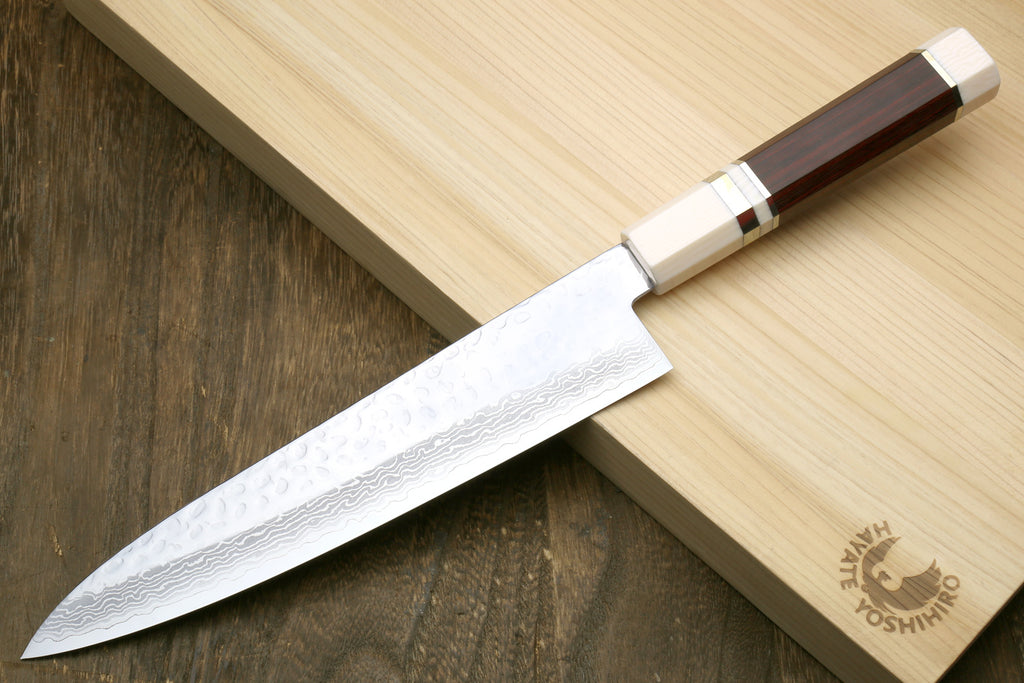 A Japanese-style kitchen knife with a hammered Damascus steel blade and a dark brown wooden handle, resting diagonally on a light wood cutting board over a dark wooden surface. The cutting board features the brand logo 'Oishihiro'.