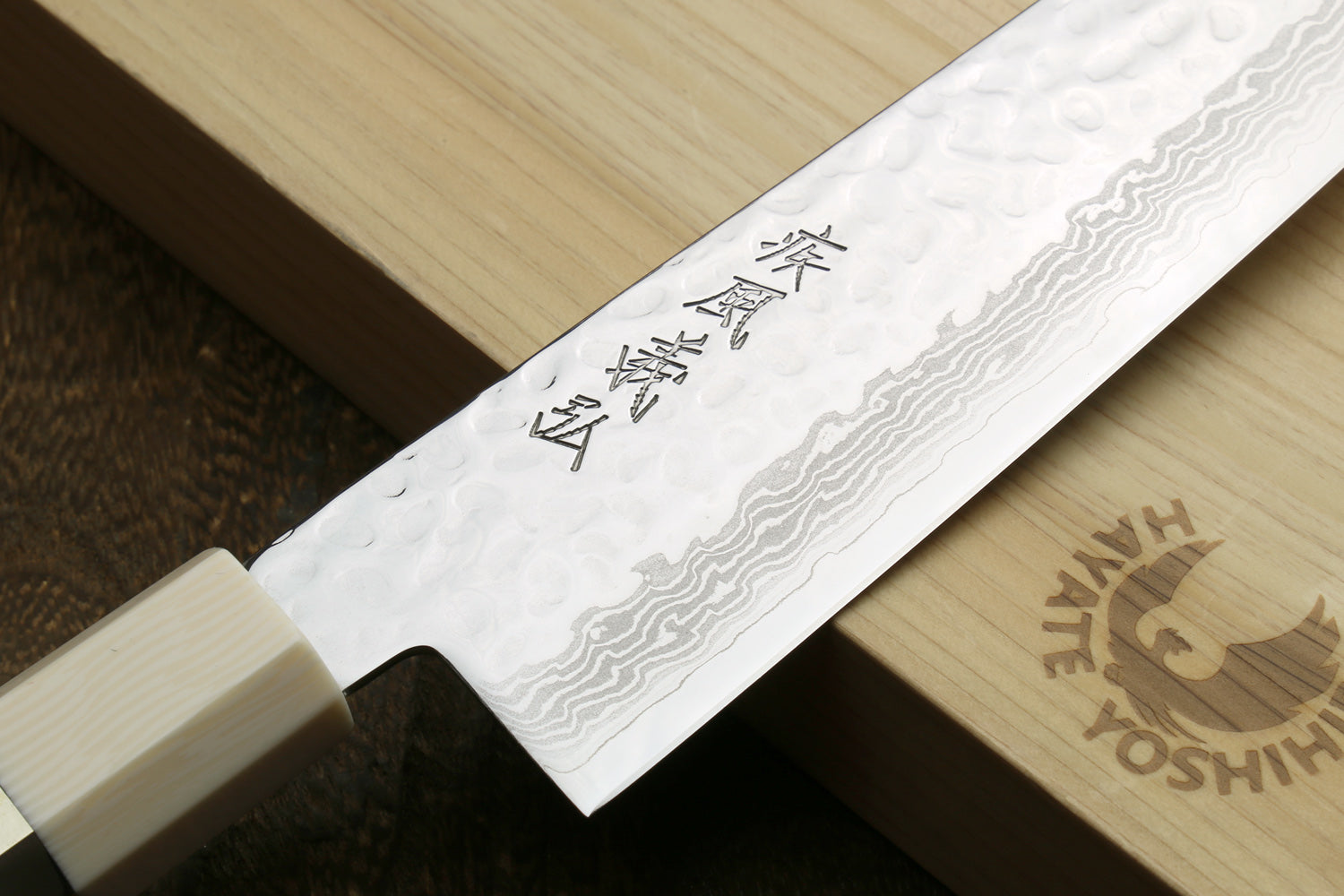 A close-up of a Japanese kitchen knife with a hammered blade finish and engraved kanji, resting on a light wood cutting board with the Haniya Yoshihira logo visible.