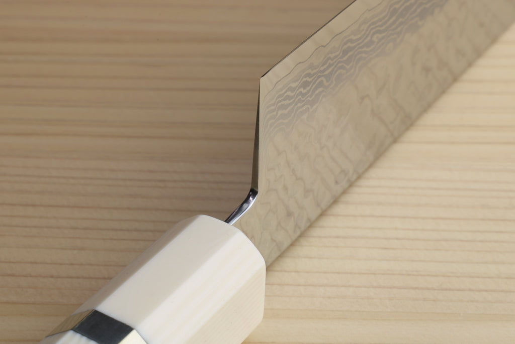 A close-up of a Japanese-style kitchen knife with a Damascus steel blade and a cream-colored handle, resting on a light wood grain surface.