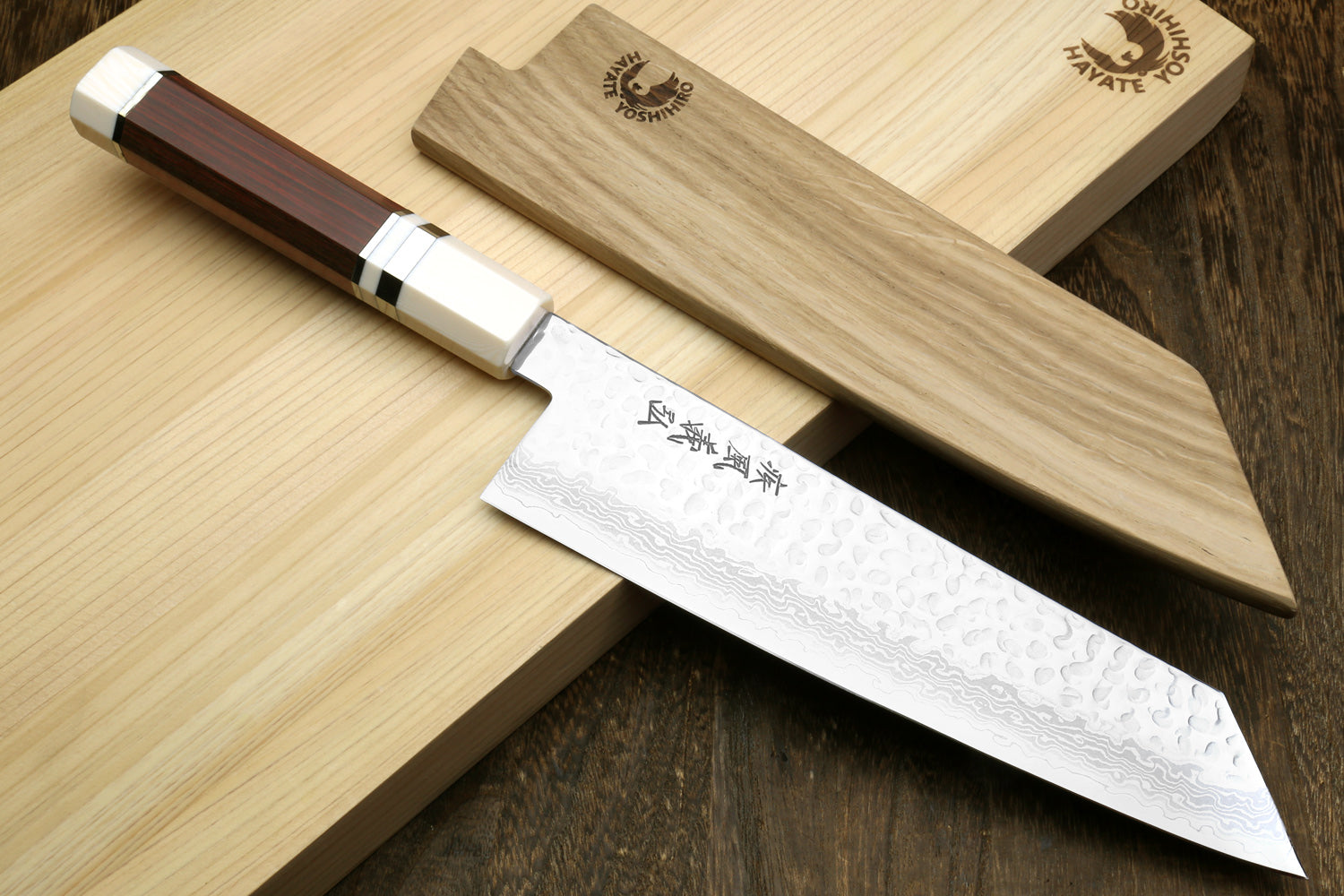 A Japanese-style kitchen knife with a hammered Damascus steel blade and a dark brown wooden handle, displayed diagonally on a light wood cutting board next to its matching wooden sheath.