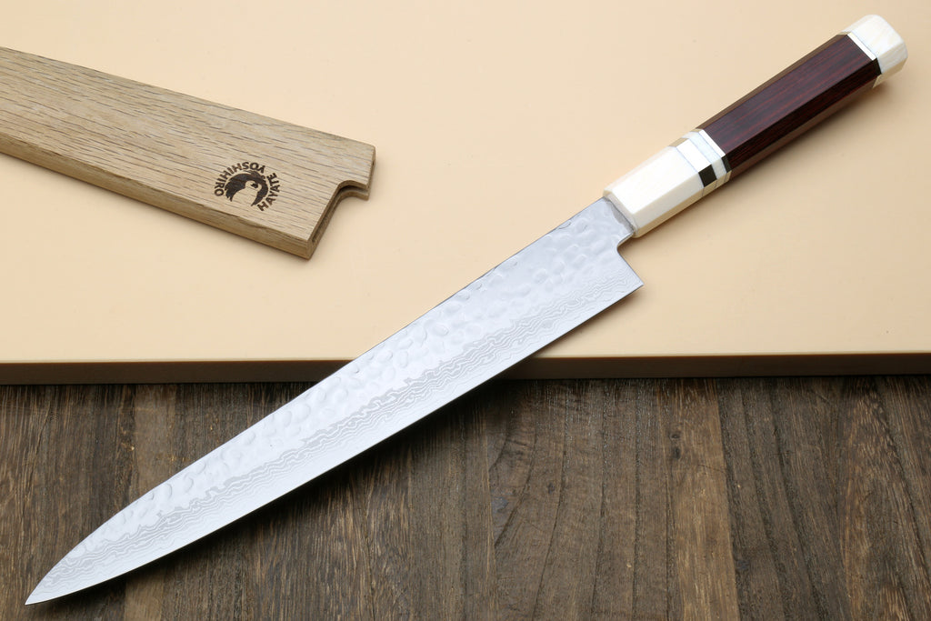 A Japanese-style kitchen knife with a hammered Damascus steel blade, a silver bolster, and a dark reddish-brown wooden handle, shown alongside its matching wooden sheath on a beige background with a dark wood surface below.