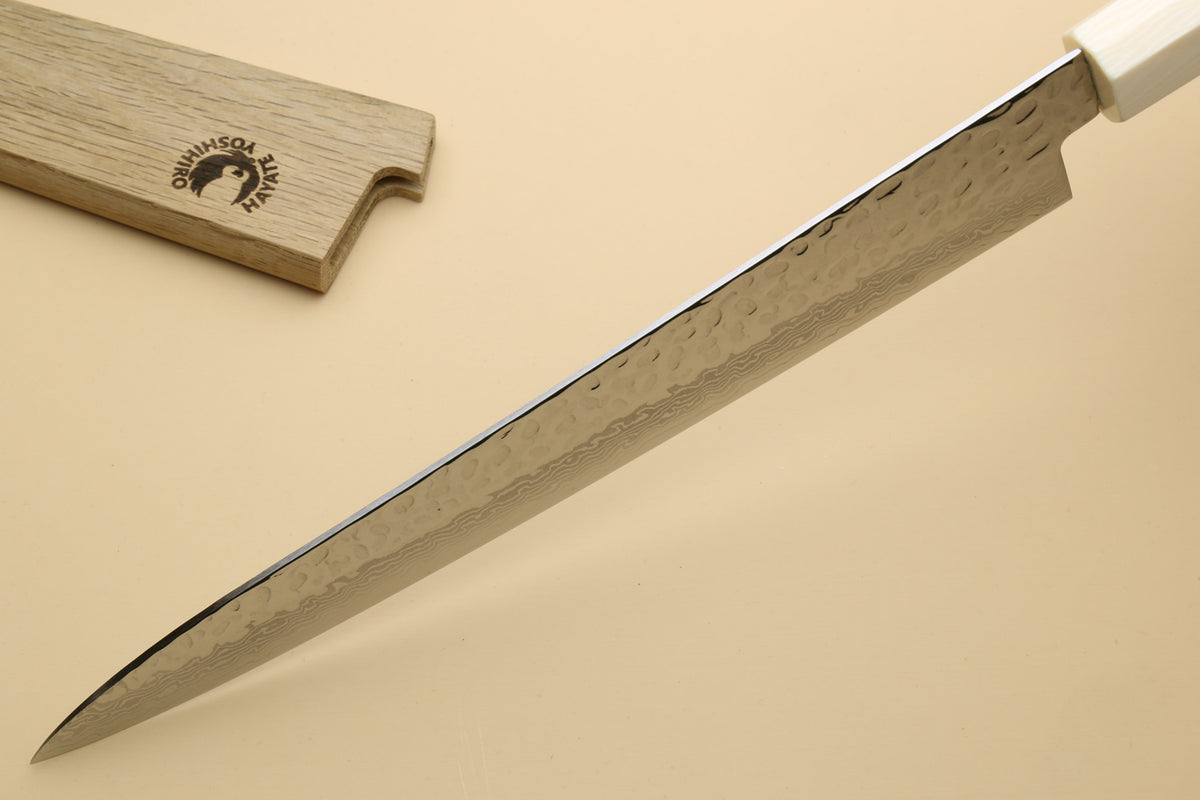 A close-up of a Japanese-style kitchen knife with a hammered Damascus steel blade and a white handle, shown diagonally on a beige background with its wooden box partially visible in the top left.