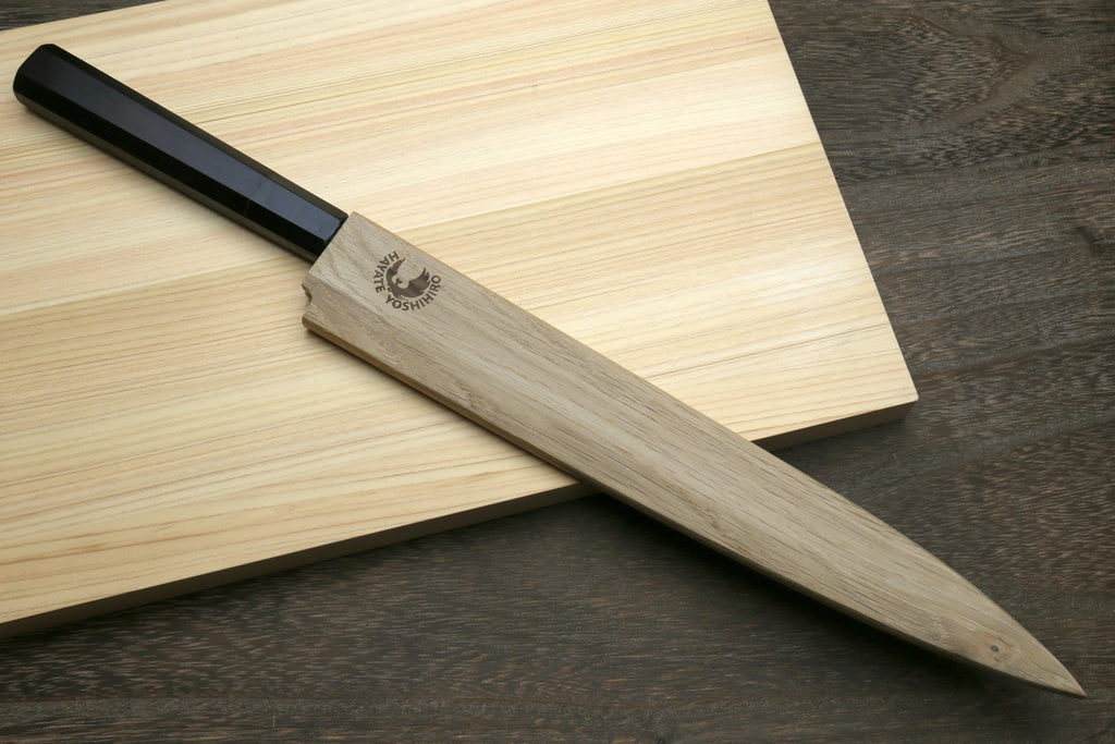 Yanagi knife with an ebony handle stored in a wooden sheath on a wooden cutting board.