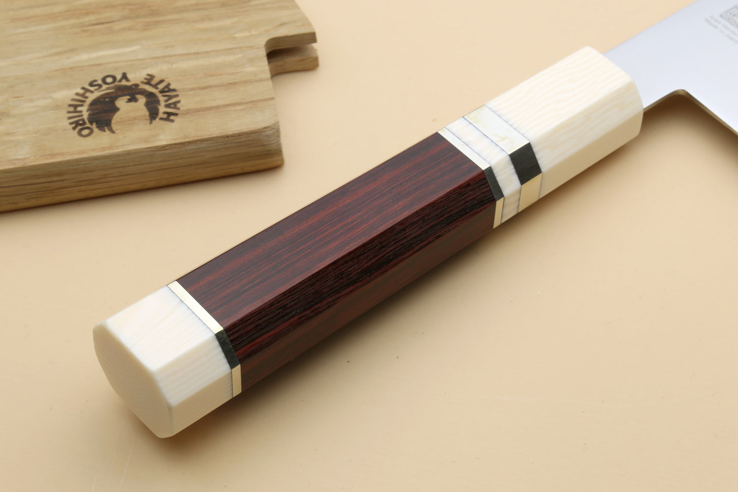 A close-up of a Japanese-style kitchen knife with a dark reddish-brown wood handle and cream-colored end caps, resting on a beige surface next to a wooden cutting board.