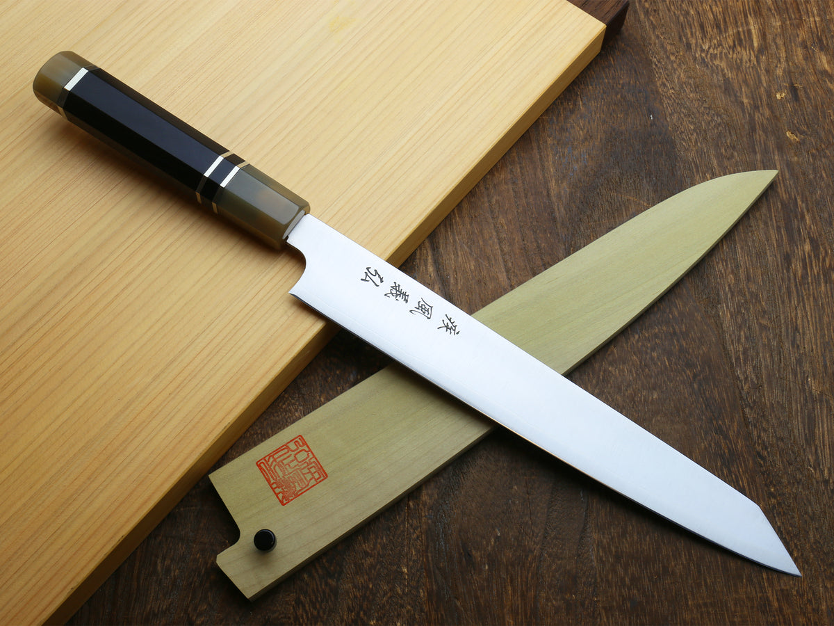 Sujihiki kiritsuke knife with white blade, dark horn handle, and wooden sheath on boards.