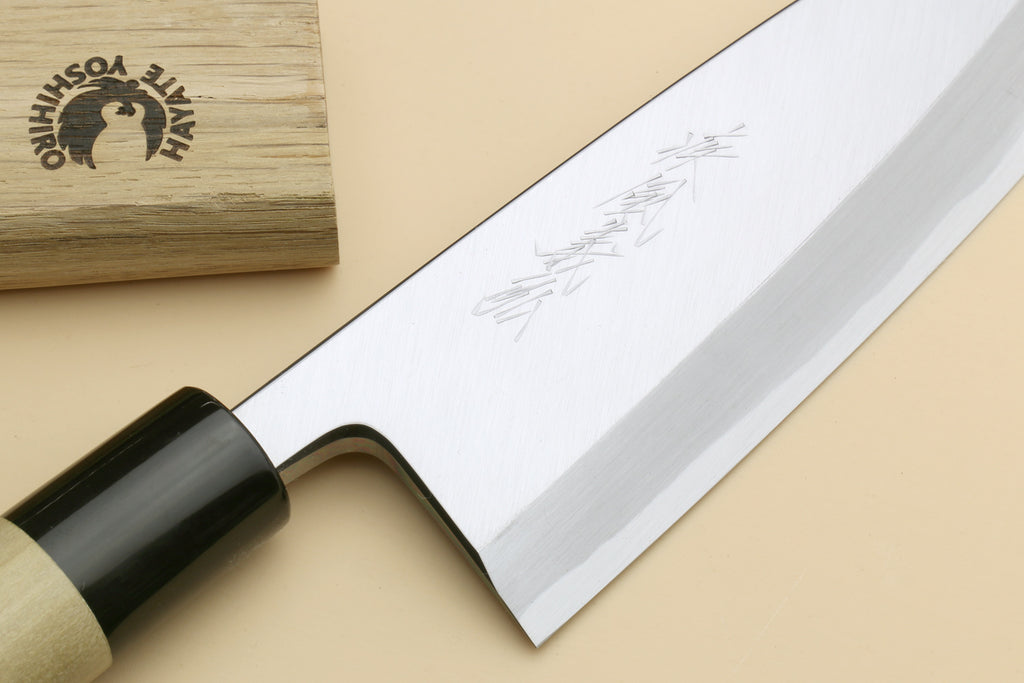 A Japanese-style kitchen knife with a polished silver blade, black ferrule, and light wood handle, shown diagonally on a beige background next to its wooden presentation box engraved with the brand 'Hate Yoshitaka'.