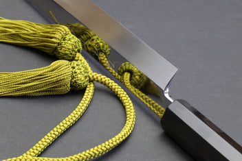 Mirror-finished chef knife blade with pointed tip and dark ebony handle, cord tied.