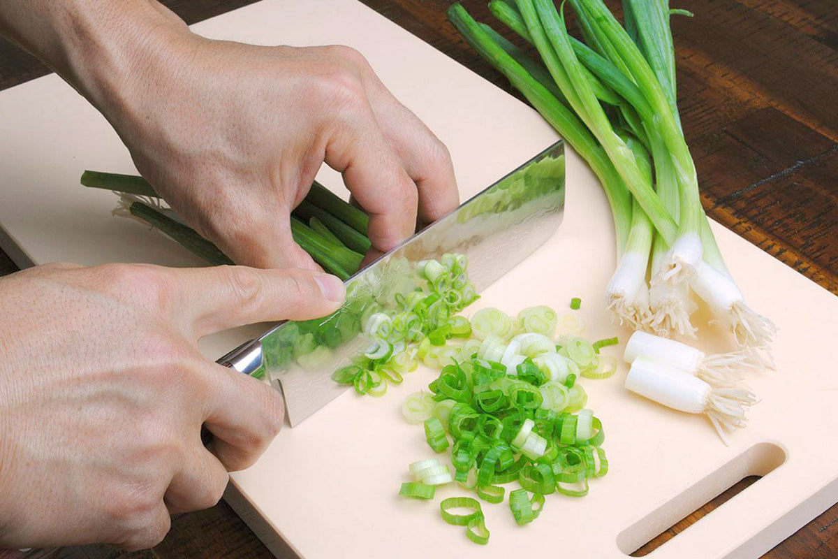Nakiri knife slicing green onions on a cutting board with dark wood handle